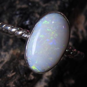 Op de afbeelding: Zilveren ring met een ovale opaal edelsteen. De opaal heeft een melkachtige witte basis met groene, blauwe en gele flitsen. De band heeft een gedraaid touwontwerp. Sieraden.