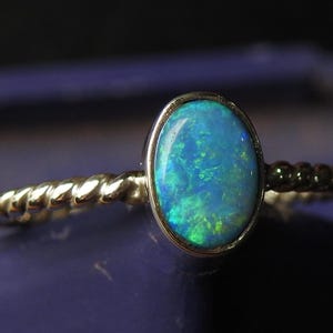 Op de afbeelding: Een goudkleurige ring met een ovale opaal edelsteen met blauwe en groene tinten. De band heeft een gedraaid touwontwerp. De opaal heeft een iriserend effect. De ring staat op een donkerblauwe achtergrond.