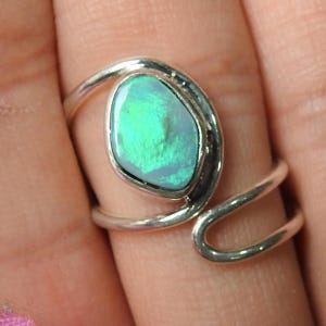 Op de afbeelding: Zilveren ring met een ovale opaal, met levendige groene en blauwe tinten. De band van de ring heeft een uniek, vloeiend ontwerp dat zich om de vinger wikkelt. De opaal is gezet in een zilveren zetting.