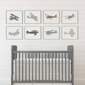 Peut inclure: Un lit b&eacute;b&eacute; gris avec un matelas blanc et une jupe &agrave; volants blanche. Le lit b&eacute;b&eacute; est devant un mur blanc avec huit impressions encadr&eacute;es de silhouettes d'avions dans diff&eacute;rentes nuances de gris, de brun et de beige.