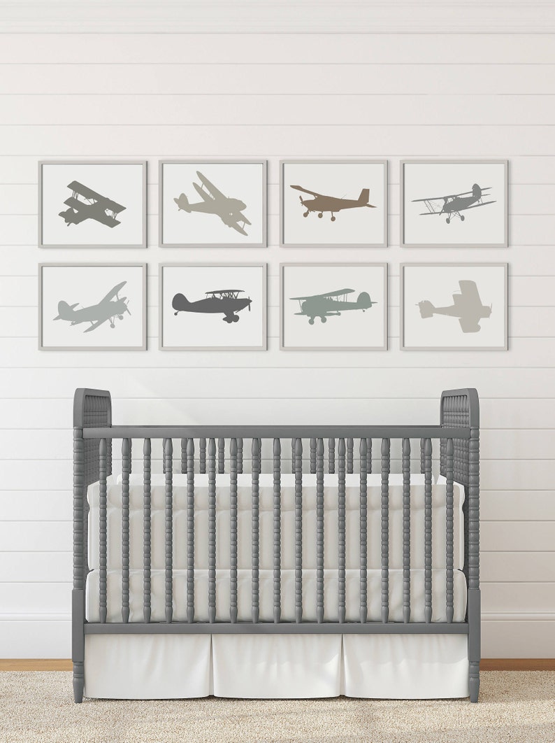 Peut inclure: Un lit b&eacute;b&eacute; gris avec un matelas blanc et une jupe &agrave; volants blanche. Le lit b&eacute;b&eacute; est devant un mur blanc avec huit impressions encadr&eacute;es de silhouettes d'avions dans diff&eacute;rentes nuances de gris, de brun et de beige.