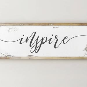 May include: White wooden sign with black calligraphy that reads "inspire" on a white background.
