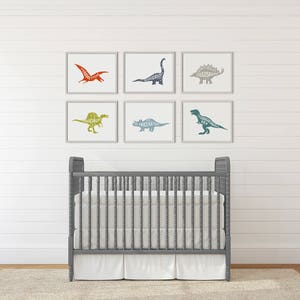 Puede incluir: Seis impresiones art&iacute;sticas de dinosaurios enmarcadas en tonos naranja, azul, verde y gris cuelgan sobre una cuna gris con un colch&oacute;n y una falda blancos. Las siluetas de los dinosaurios est&aacute;n rellenas con el nombre del dinosaurio. La cuna est&aacute; sobre una alfombra beige.