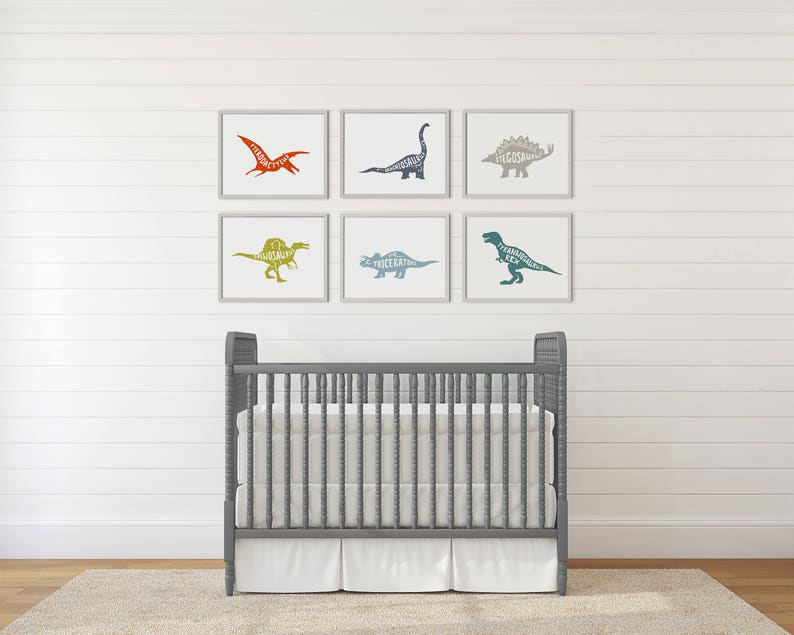 Puede incluir: Seis impresiones art&iacute;sticas de dinosaurios enmarcadas en tonos naranja, azul, verde y gris cuelgan sobre una cuna gris con un colch&oacute;n y una falda blancos. Las siluetas de los dinosaurios est&aacute;n rellenas con el nombre del dinosaurio. La cuna est&aacute; sobre una alfombra beige.