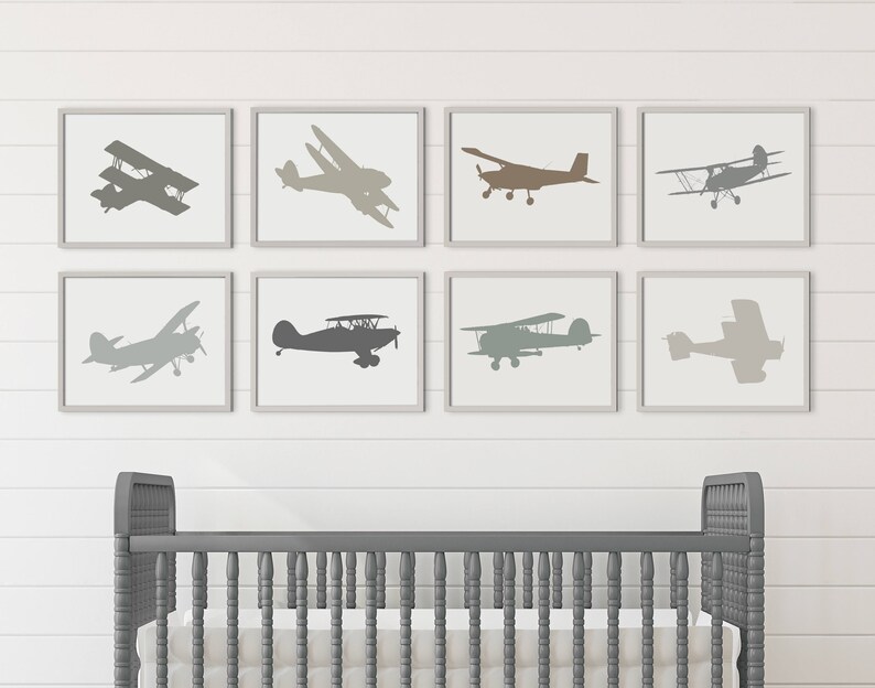 Peut inclure: Un ensemble de huit impressions encadr&eacute;es repr&eacute;sentant des silhouettes d'avions dans diff&eacute;rentes nuances de gris. Les impressions sont dispos&eacute;es en grille sur un mur blanc au-dessus d'un berceau gris.