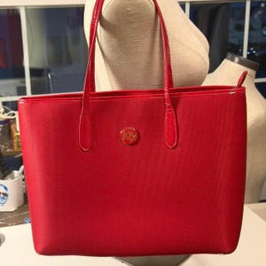May include: A bright red tote bag with a textured fabric exterior and two matching red shoulder straps. The front of the bag features a circular gold-toned metal emblem in the center with the word JOY embossed in the middle of the hardware.