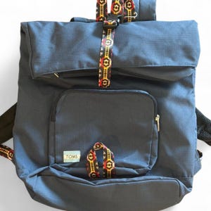 May include: A navy blue backpack featuring a roll-top closure secured by a black plastic buckle on a patterned strap with red, yellow, and black geometric designs. A front zippered pocket displays a light blue TOMS label and a matching patterned loop.