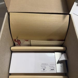 May include: A light-colored wood memo pad holder with a curved top piece and a flat base, packaged inside a cardboard box. The set includes a white paper refill pad with the text DOODAD and instructions printed on the label, plus a metal pin.