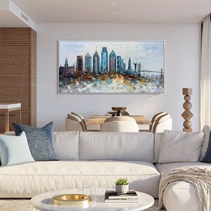 Pode incluir: Um sof&aacute; seccional branco com almofadas azuis em uma sala de estar moderna. Uma grande pintura abstrata de um horizonte da cidade fica pendurada acima do sof&aacute;. A pintura apresenta uma ponte e edif&iacute;cios em tons de azul, marrom e branco.