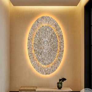 Op de afbeelding: Een groot ovaal wandkunstwerk met een radiaal patroon van dunne, lichtgekleurde, getextureerde stroken in concentrische cirkels. Het kunstwerk is warm verlicht van achteren, wat een halo-effect creëert. Een kleine vaas staat eronder.
