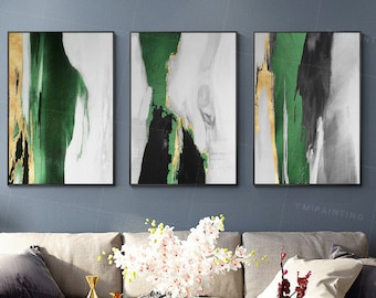 May include: Three abstract paintings in a modern living room. The paintings are in shades of green, black, and gold. The paintings are hung on a gray wall above a beige sofa. The sofa is decorated with throw pillows and a vase of white flowers.