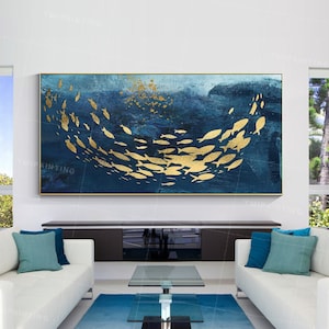 May include: A large abstract painting of a school of gold fish swimming in a blue ocean. The painting is framed in gold and hangs on a white wall above a white couch and a glass coffee table.