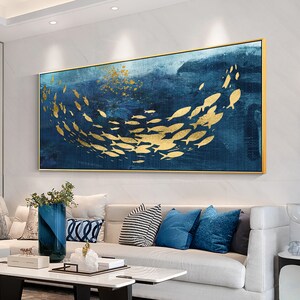 May include: Abstract painting of a school of gold fish swimming in a blue ocean. The painting is framed in a gold frame and is hanging on a white wall.