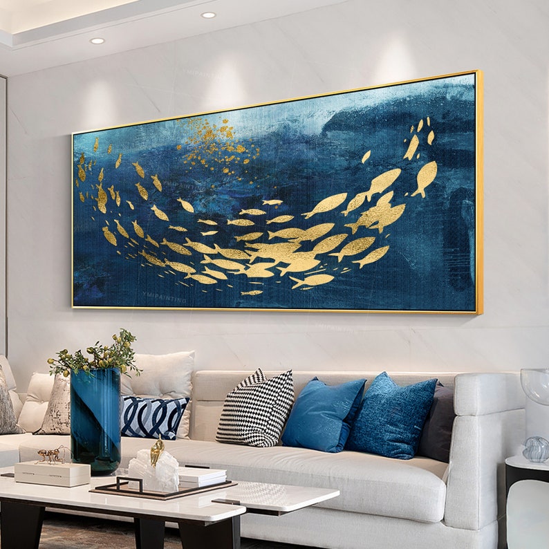 May include: Abstract painting of a school of gold fish swimming in a blue ocean. The painting is framed in a gold frame and is hanging on a white wall.