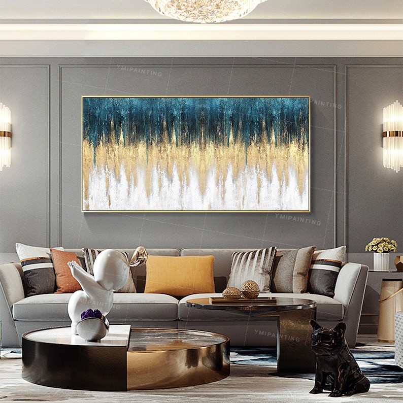 Op de afbeelding: Abstract schilderij in blauw, goud en wit in een horizontaal formaat. Het schilderij is ingelijst in goud en hangt aan een grijze muur boven een grijze bank met kussens.