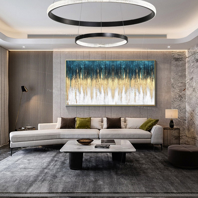 May include: Abstract painting with blue, gold, and white colors. The painting is framed in a gold frame and is hanging on a wall in a living room. The painting is above a white couch with green pillows.