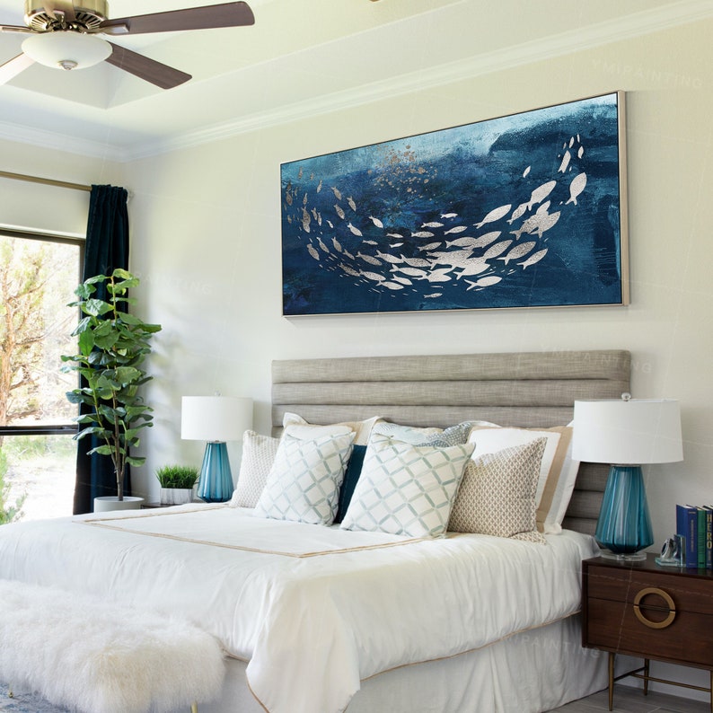 May include: A white bed with a gray headboard and a blue and silver abstract painting of fish above it. The bed has a white duvet and several pillows, including a blue and white patterned pillow.