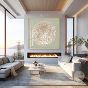 Op de afbeelding: Een moderne woonkamer met een groot, vierkant kunstwerk als middelpunt. Het kunstwerk heeft een cirkelvormig, gestructureerd ontwerp in crème- en goudtinten, tegen een lichtgroene achtergrond. De kamer omvat een open haard, banken en grote ramen.