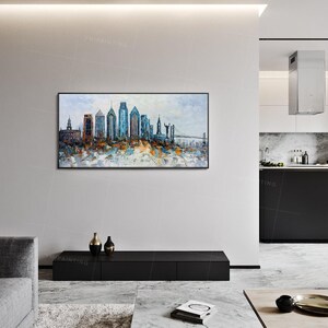 Pode incluir: Uma pintura de um horizonte urbano com edif&iacute;cios altos em tons de azul, marrom e branco. A pintura &eacute; emoldurada e pendurada em uma parede branca em uma sala de estar moderna.