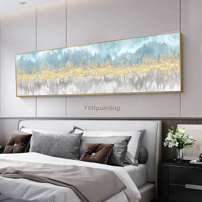 May include: A long, horizontal abstract painting with a gold frame. The painting features a blue and gray background with gold accents. The painting is hanging on a wall above a bed with gray pillows.