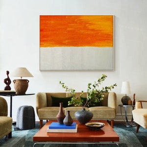 Puede incluir: Pintura abstracta con una banda horizontal de pintura naranja sobre una banda blanca. La pintura est&aacute; colgada en una pared blanca sobre un sof&aacute; marr&oacute;n con plantas verdes y jarrones marrones.