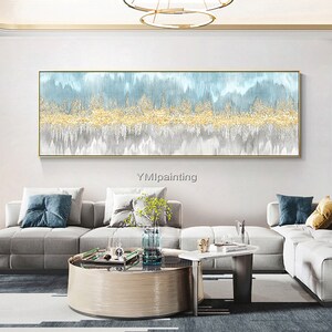 May include: Abstract painting with a blue, gold, and gray color scheme. The painting features a horizontal band of gold paint with a textured effect, surrounded by blue and gray washes. The painting is framed in a thin gold frame and is hanging on a white wall in a living room setting.