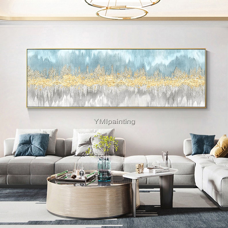 May include: Abstract painting with a blue, gold, and gray color scheme. The painting features a horizontal band of gold paint with a textured effect, surrounded by blue and gray washes. The painting is framed in a thin gold frame and is hanging on a white wall in a living room setting.