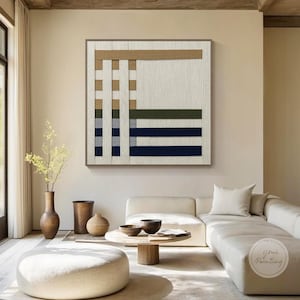 Puede incluir: Una gran obra de arte mural abstracta cuadrada con una textura tejida. El diseño presenta bandas horizontales y verticales en tonos beige, marrón, azul marino y verde oliva. La obra está enmarcada en madera marrón claro y cuelga sobre un sofá blanco.