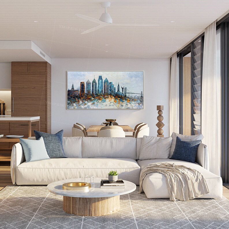 Pode incluir: Um sof&aacute; seccional branco com almofadas azuis em uma sala de estar moderna. Uma grande pintura abstrata de um horizonte da cidade fica pendurada acima do sof&aacute;. A pintura apresenta uma ponte e edif&iacute;cios em tons de azul, marrom e branco.