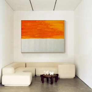 Puede incluir: Un sof&aacute; seccional blanco con una mesa de caf&eacute; de madera y tres objetos decorativos blancos en la parte superior. Una gran pintura abstracta con rayas naranjas y blancas cuelga de la pared detr&aacute;s del sof&aacute;.