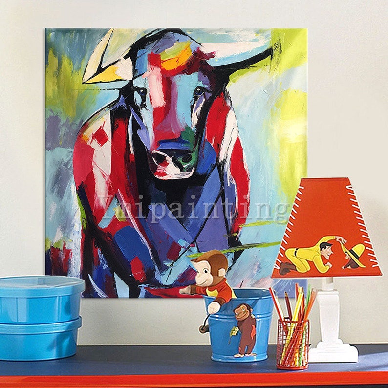 May include: Abstract painting of a colorful bull with red, blue, yellow, and white paint. The bull is facing forward with its horns pointing up. The painting is on a canvas and is framed.