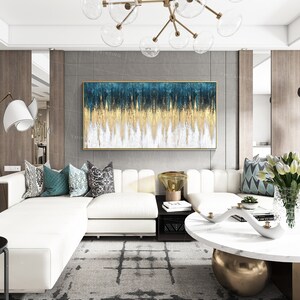 Op de afbeelding: Abstract schilderij in blauw, goud en wit. Het schilderij is ingelijst en hangt aan een grijze muur boven een witte hoekbank.