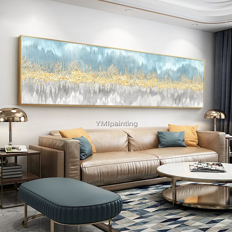 May include: A large abstract painting with a gold, blue, and gray color scheme. The painting is framed in a gold frame and is hanging on a wall in a living room. The painting is above a brown leather sofa with blue and yellow throw pillows. A round coffee table with a glass top is in front of the sofa. The coffee table is sitting on a patterned rug.