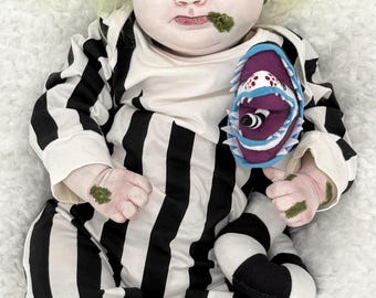 May include: A realistic baby doll with pale skin, green hair, and moss-like patches on the face, hands, and feet. The doll wears a black and white striped long-sleeve romper and holds a small purple, blue, and white creature plush toy.
