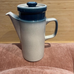 May include: A ceramic coffee pot with a blue and white speckled design. The pot has a handle, a spout, and a lid with a blue knob. The pot is sitting on a pink surface and against a wooden background.