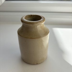 Può includere: Un piccolo vaso antico in gres, con corpo cilindrico e spalla arrotondata. Il vaso è di colore beige chiaro con una fascia più scura nella parte superiore. L'apertura è piccola e circolare. Probabilmente usato per la conservazione.