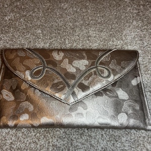 May include: A metallic silver clutch with a camouflage pattern. The flap features decorative stitching in a looping design. The clutch is rectangular and appears to be made of a leather-like material.