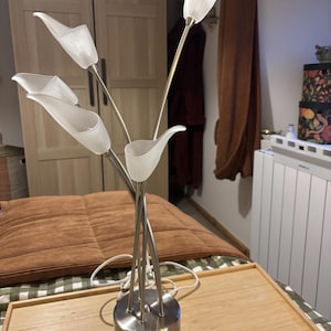 Puede incluir: Lámpara de mesa decorativa con diseño floral. Presenta cinco pantallas blancas en forma de tulipán sobre finos tallos plateados. La base es un cilindro de metal cepillado. La lámpara está sobre una bandeja de madera.