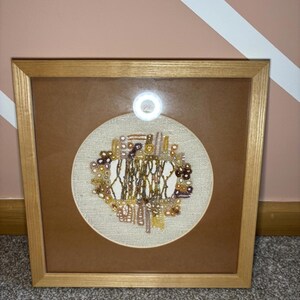 May include: Framed artwork with an embroidered design in brown, beige, and gold. The embroidery is set against a beige fabric background, framed in a light brown wooden frame. The artwork is displayed on a grey carpet.