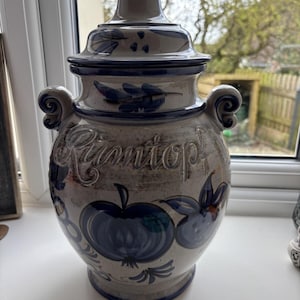 May include: A ceramic jar with a lid, in grey and blue tones. The jar is decorated with blue apple motifs and the word "Rumtopf" is embossed on the front. It has two ornate handles. The jar is sitting on a white surface.