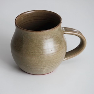 May include: A handmade ceramic mug with a rounded body and a wide, slightly flared rim. The mug is a muted green colour with a subtle brown glaze and a red rim.