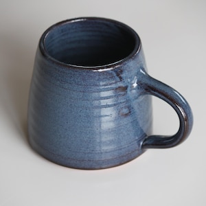 May include: A handmade blue ceramic mug with a rounded body and a curved handle. The mug has a textured surface and a dark blue glaze, perfect for coffee or tea. The mug is approximately 10 cm tall.