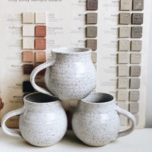 Peut inclure: Trois tasses en céramique blanche avec un glaçage moucheté empilées les unes sur les autres. Les tasses ont un corps arrondi et une anse. Les tasses sont devant un tableau avec des échantillons de couleurs.