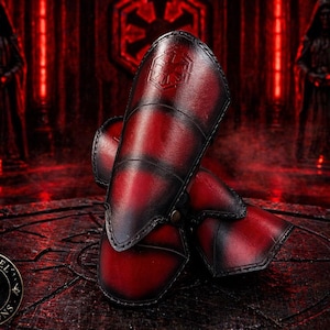 Könnte beinhalten: Rote und schwarze Leder-Armschützer mit einem genähten Design und einem Metallverschluss. Die Armschützer haben ein geprägtes Symbol. Der Hintergrund ist eine dunkelrote Szene mit Figuren und einem Symbol.