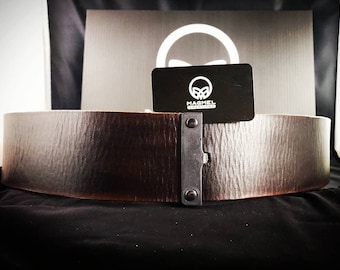 May include: A brown leather belt with a black metal buckle. The buckle has a small metal plate with the text "MAGMEL CREATIONS" on it.