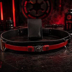 May include: A black utility belt featuring a prominent red leather strap overlay, a central metallic buckle with a circular emblem, and a vertical black pouch attached to the rear. The belt includes silver-toned hardware and small black symbols.