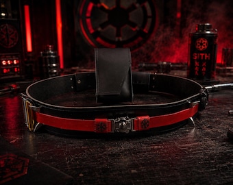 May include: A black utility belt featuring a prominent red leather strap overlay, a central metallic buckle with a circular emblem, and a vertical black pouch attached to the rear. The belt includes silver-toned hardware and small black symbols.