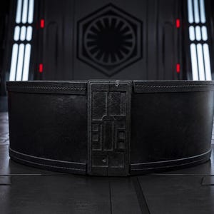 Puede incluir: Un cinturón ancho de cuero negro con una hebilla rectangular con un diseño geométrico. El cinturón tiene un borde cosido. El fondo incluye cascos de Stormtroopers y un interior futurista.