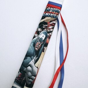 Può includere: Una candela bianca con un nastro rosso, bianco e blu. La candela è decorata con un'immagine a fumetti di Capitan America. Il testo "Captain America" è visibile sulla candela.