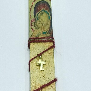 Può includere: Una candela religiosa bianca e dorata con un'immagine rossa e blu della Vergine Maria e del bambino Gesù. La candela è avvolta da uno spago rosso e dorato e ha un ciondolo a forma di croce dorata.